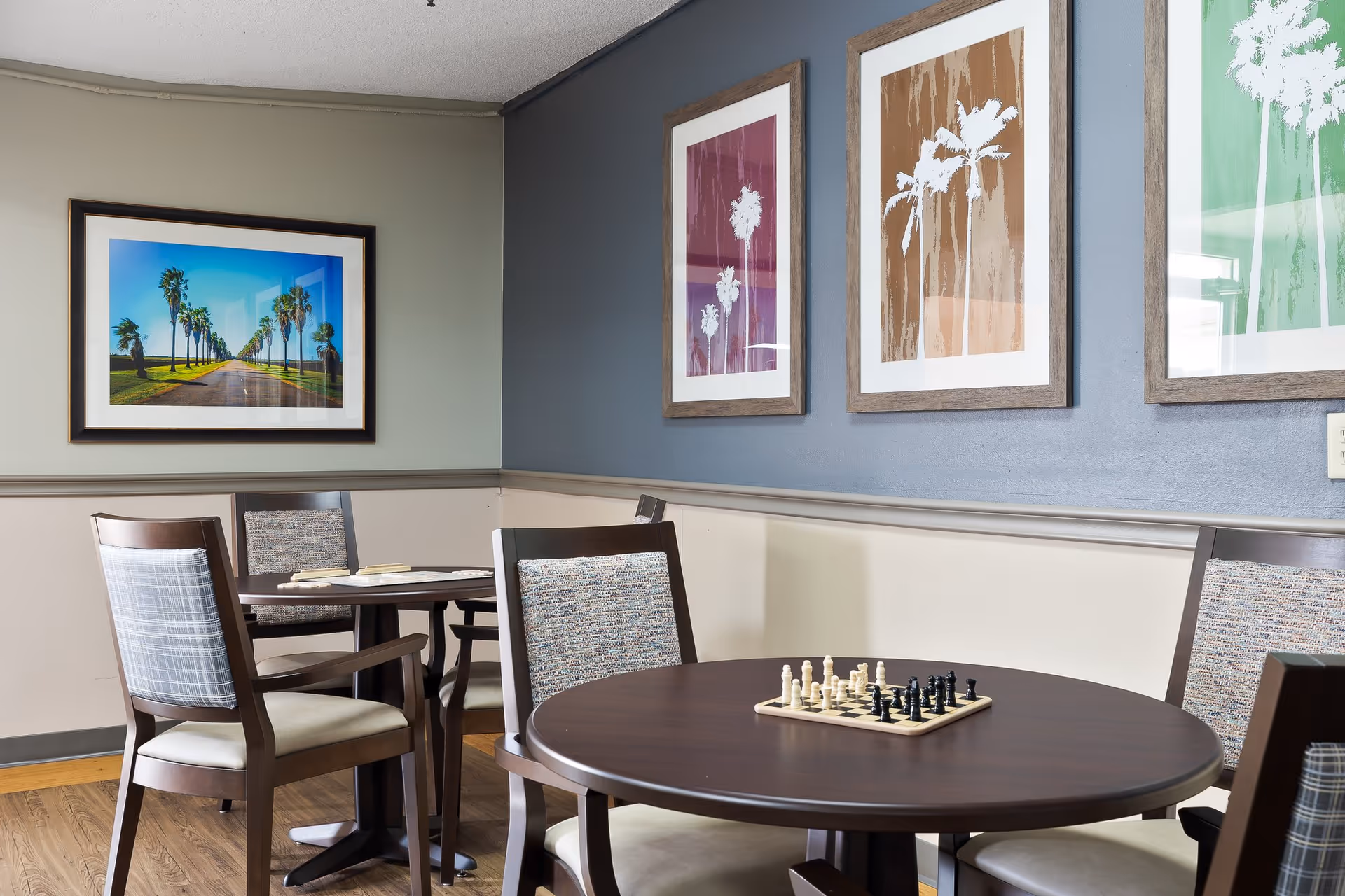 A cozy common area with round wooden tables and cushioned chairs. One table has a chessboard set up with chess pieces. The walls are decorated with framed artwork featuring palm trees in various colors. The floor is wooden, and the room has a calm, inviting atmosphere.