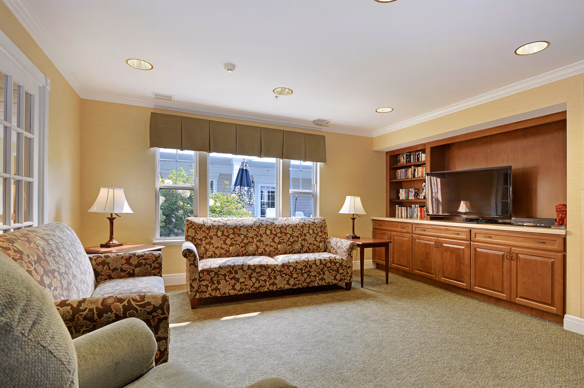 Cozy communal living room with patterned sofas, side tables and lamps, a TV set in wooden built-in cabinets, and a large window.