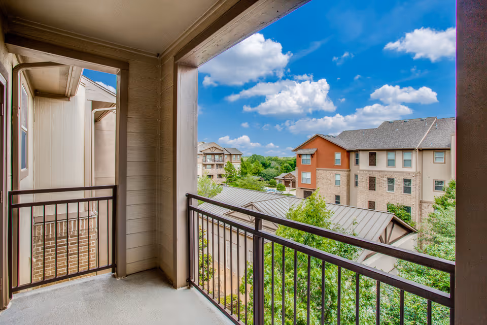 Covered second-story balcony with metal railings overlooking neighboring apartment buildings and trees under a partly cloudy sky.