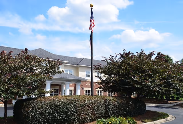 Exterior front of a senior living facility with a flagpole, columned entrance, and landscaped shrubs.