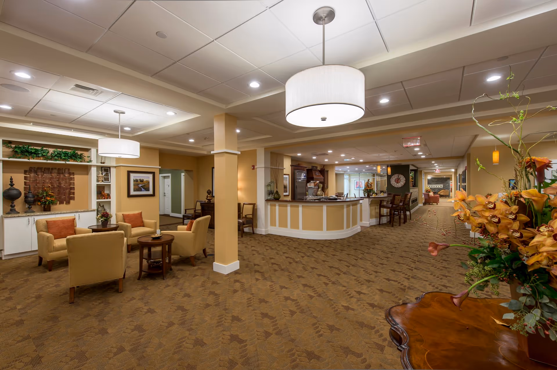 A spacious and well-lit senior living facility common area with beige walls and carpeted floors. The room features a seating area with four beige armchairs around a small wooden table, decorative plants, and artwork on the walls. There is a reception or service counter with bar stools, pendant lighting, and a large round ceiling light fixture. A floral arrangement is visible on a wooden table in the foreground.