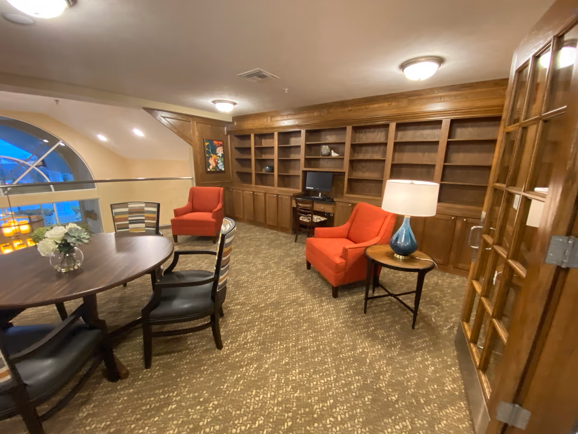 Cozy communal lounge with a round table and chairs, two red armchairs, built-in wooden bookshelves and a lamp.