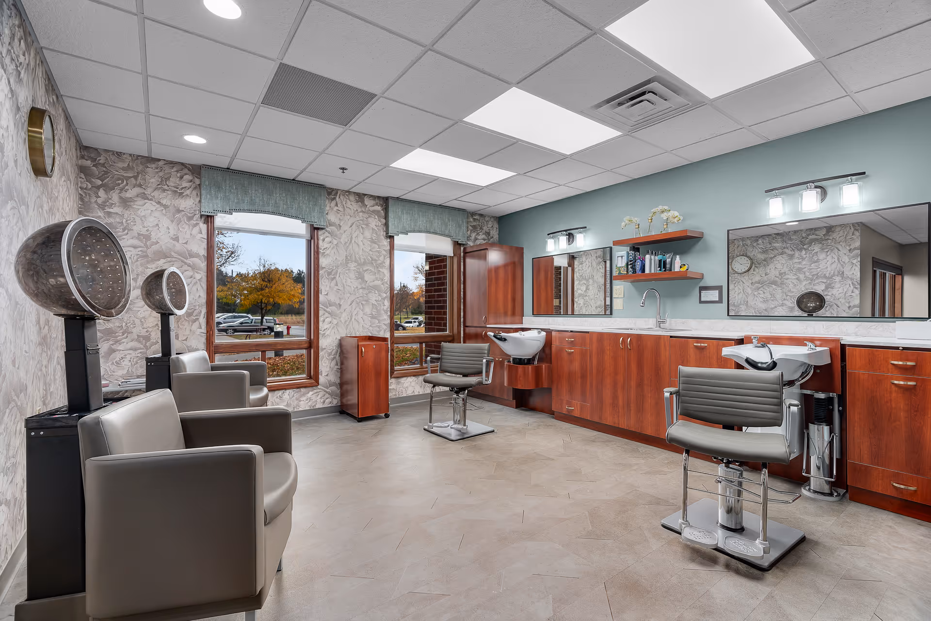 Interior view of a salon area in a senior living facility with two hair washing stations, two salon chairs, two hair dryers, large mirrors, wooden cabinets, and windows showing an outdoor view with trees and parked cars.