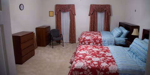 A bedroom with two twin beds, each with a red floral bedspread and blue striped pillows and sheets. The room has beige carpet, two windows with red patterned curtains, a wooden dresser, a small wooden cabinet, a chair, and a nightstand with a lamp.