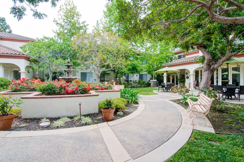 Outdoor courtyard area at a senior living facility with a curved paved walkway, benches, potted plants, a multi-tiered fountain surrounded by flowers, large trees providing shade, and a building with arched doorways and patio seating in the background.
