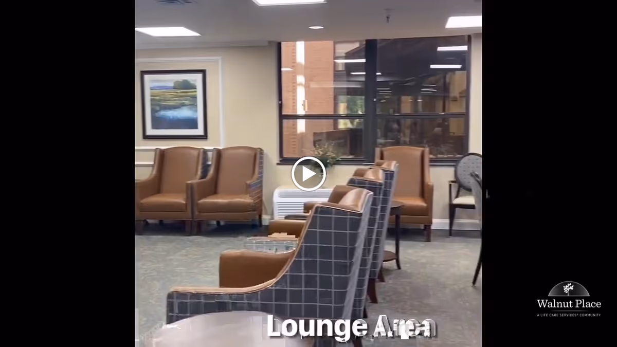A lounge area with multiple armchairs arranged in rows facing each other, a window showing a brick building outside, a framed landscape painting on the wall, and a small table with a plant. The room has carpeted flooring and neutral-colored walls.