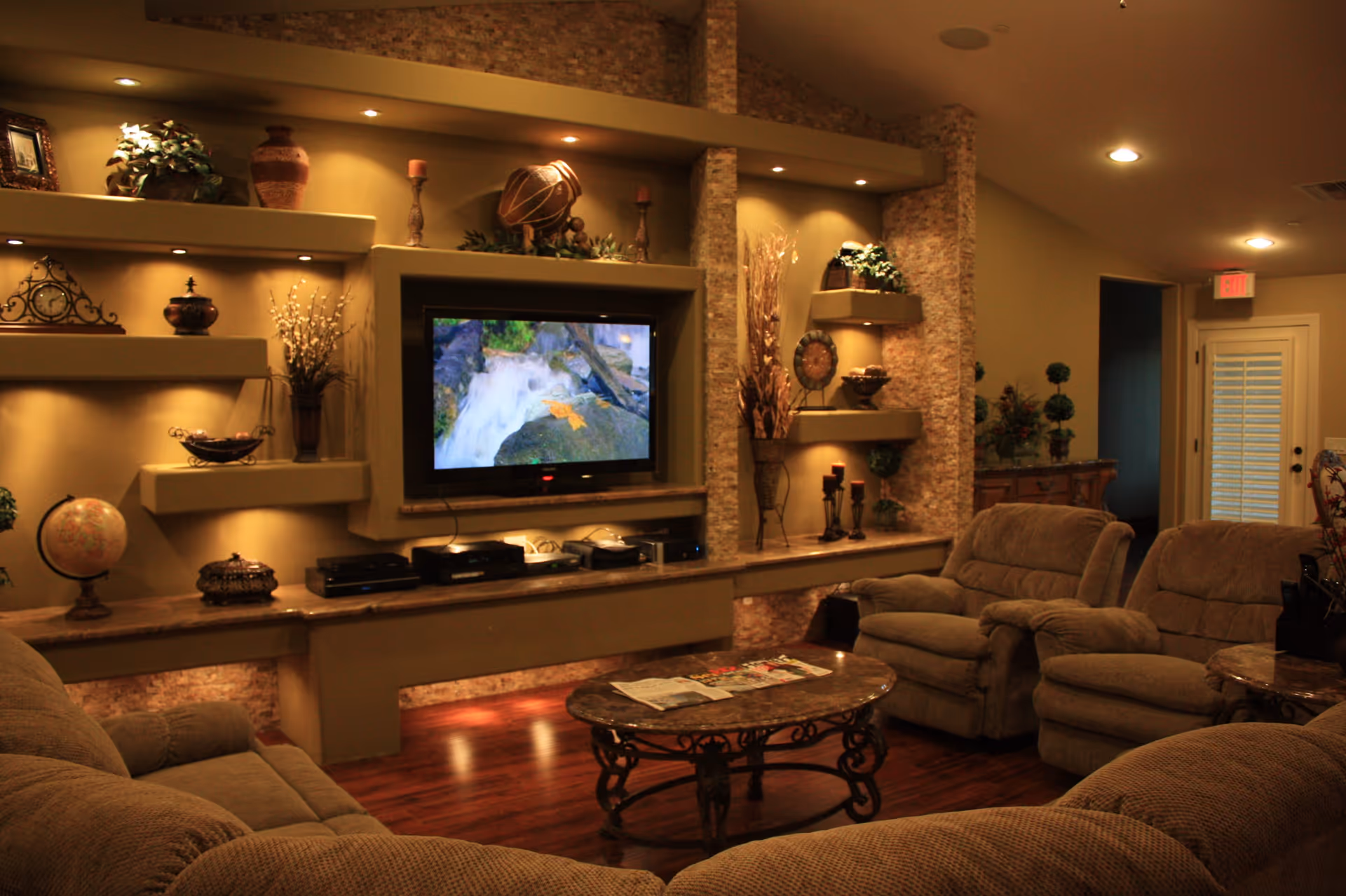 A cozy living room with plush beige sofas and armchairs arranged around a decorative coffee table. The wall features built-in shelves with various decorative items and a flat-screen TV displaying a nature scene. The room has warm lighting and wooden flooring, creating a comfortable and inviting atmosphere.