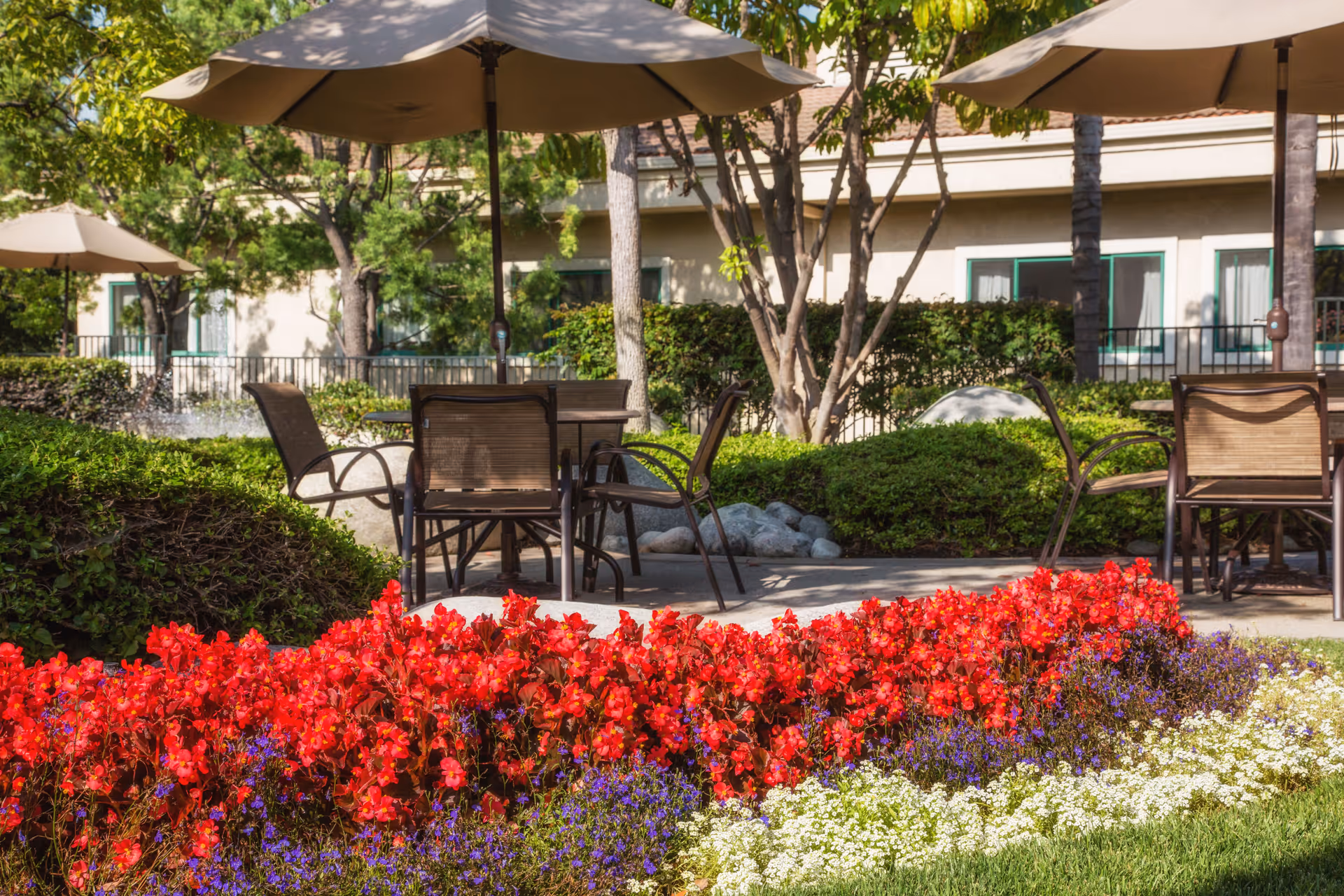 Outdoor patio area with tables and chairs under large beige umbrellas, surrounded by green bushes and colorful flower beds with red, purple, and white flowers. Trees and a building with windows are visible in the background.