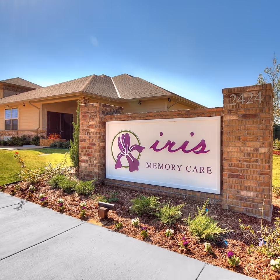 Exterior view of a brick building with a large sign in front that reads 'iris MEMORY CARE' with a purple iris flower logo. The building has a sloped roof and is surrounded by a landscaped garden with small plants and flowers. The address number 2424 is displayed on the brick pillar next to the sign.