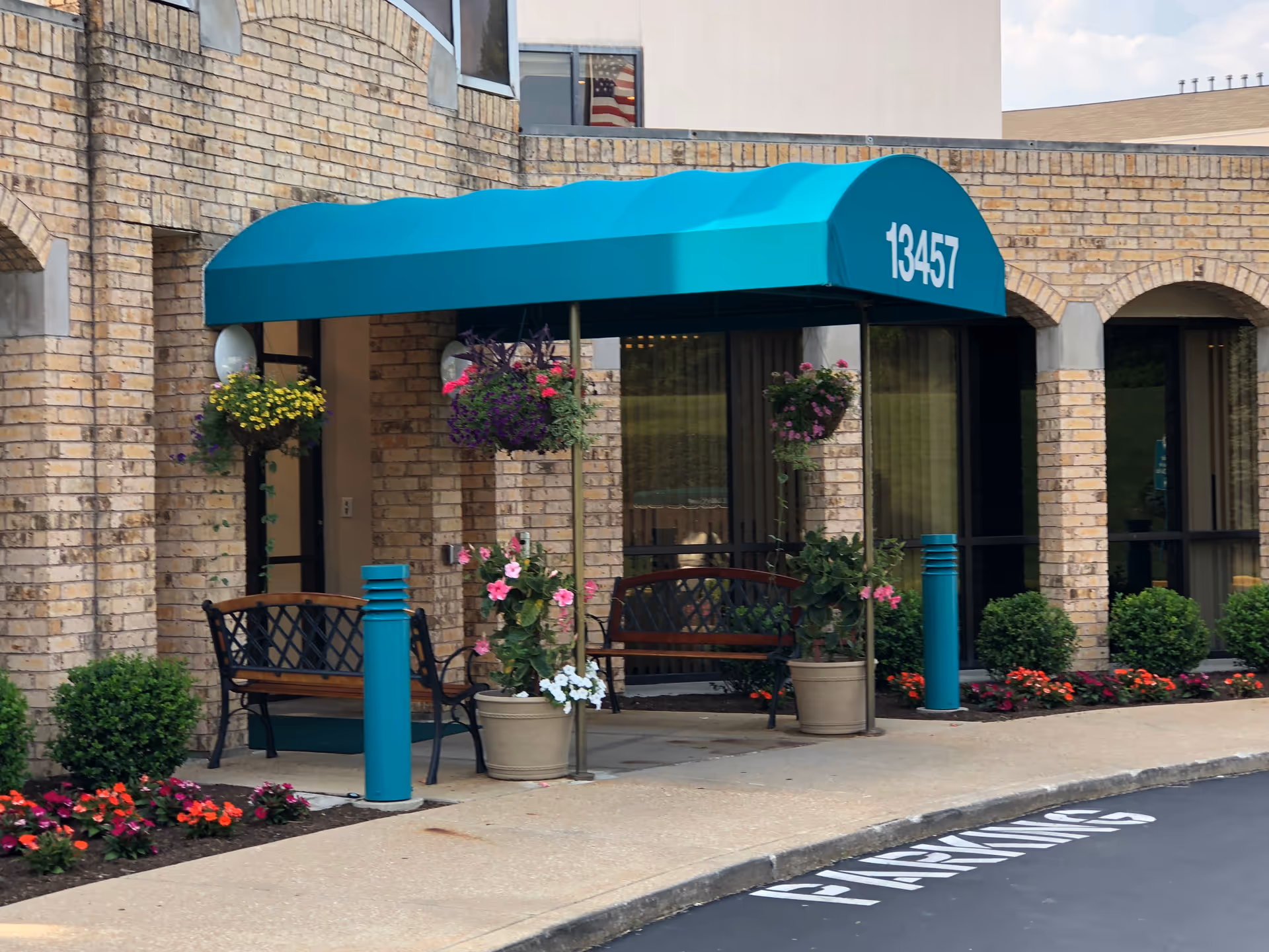 Entrance of a brick senior living building with a teal awning labeled 13457, benches, potted plants, and hanging flower baskets.
