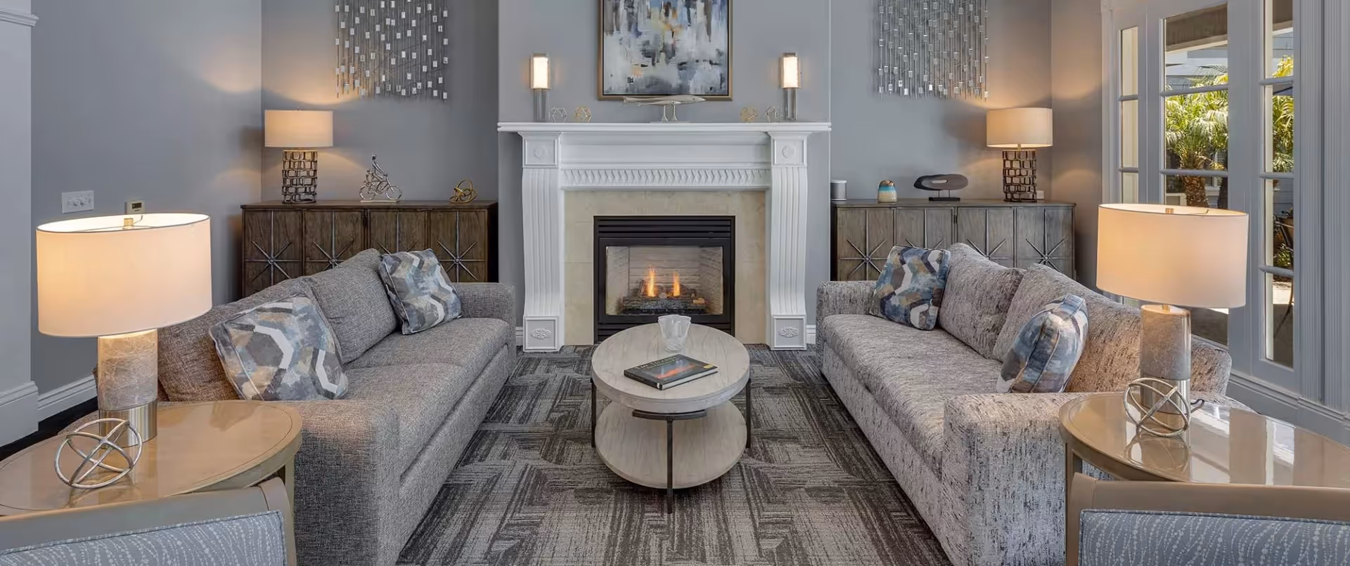 A cozy living room with two gray sofas facing each other, each adorned with patterned throw pillows. Between the sofas is a round coffee table with a book and a decorative item. The room features a white fireplace with a fire burning, flanked by two wooden cabinets topped with lamps and decorative pieces. Above the fireplace hangs an abstract painting, and the walls are painted light gray. Large windows on the right side let in natural light and show greenery outside.