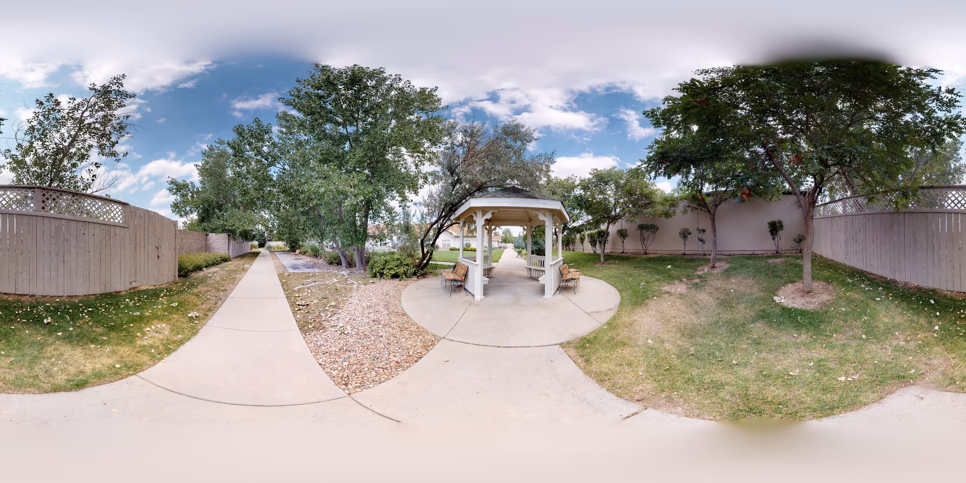 Outdoor garden area with a concrete pathway leading to a white gazebo with benches. The area is surrounded by trees, grass, and wooden fences under a partly cloudy sky.