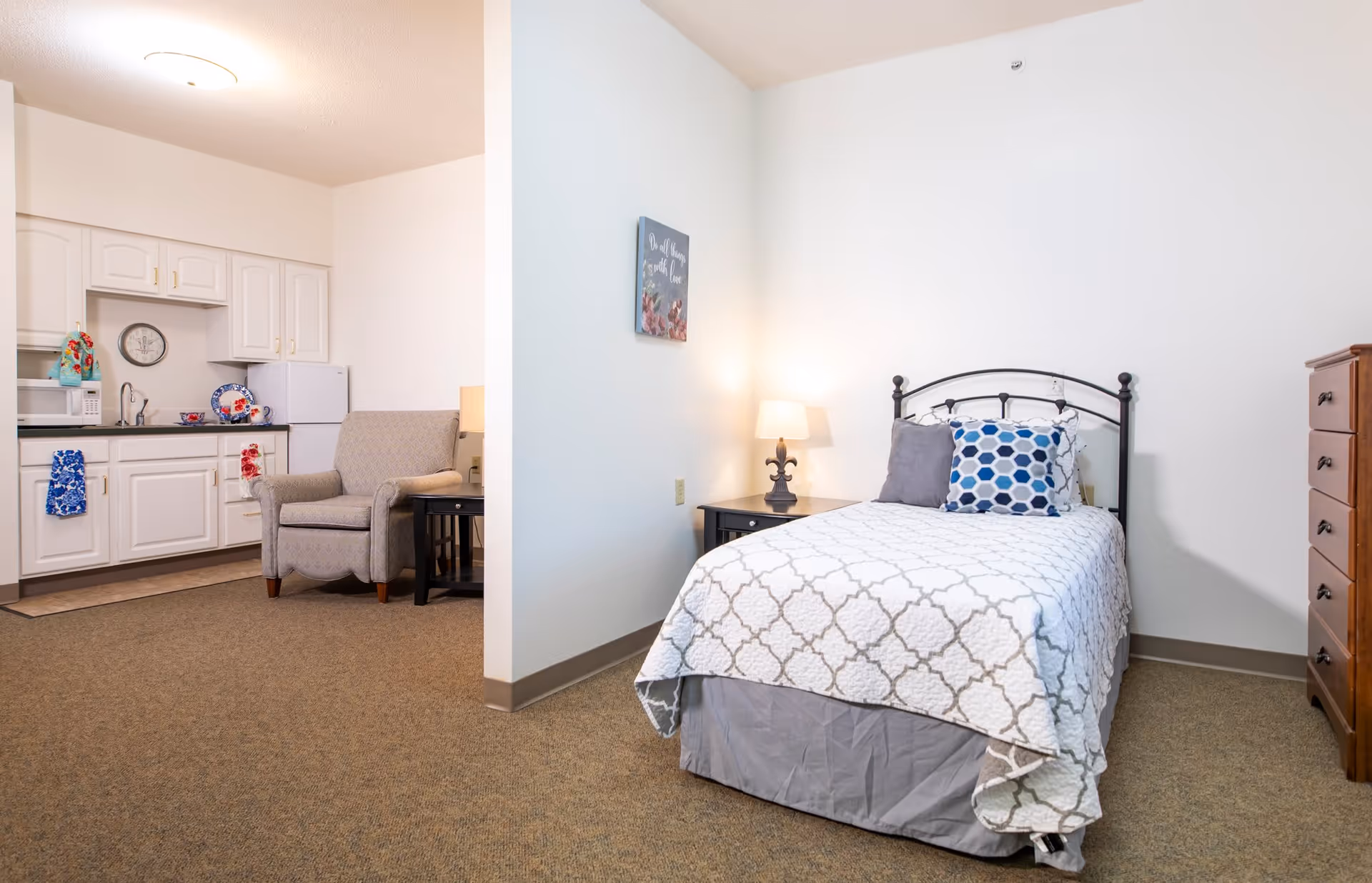 A small assisted living apartment room featuring a single bed with patterned bedding and two pillows, a nightstand with a lamp, a wooden dresser, and a wall decoration. Adjacent to the sleeping area is a kitchenette with white cabinets, a microwave, a refrigerator, a sink, and a comfortable armchair with a side table.
