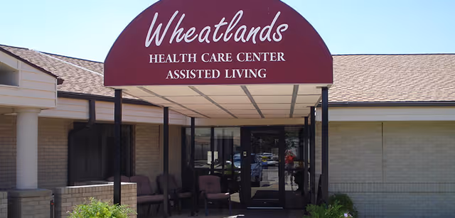 Entrance of Wheatlands Health Care Center Assisted Living with a maroon awning over glass doors and outdoor seating.