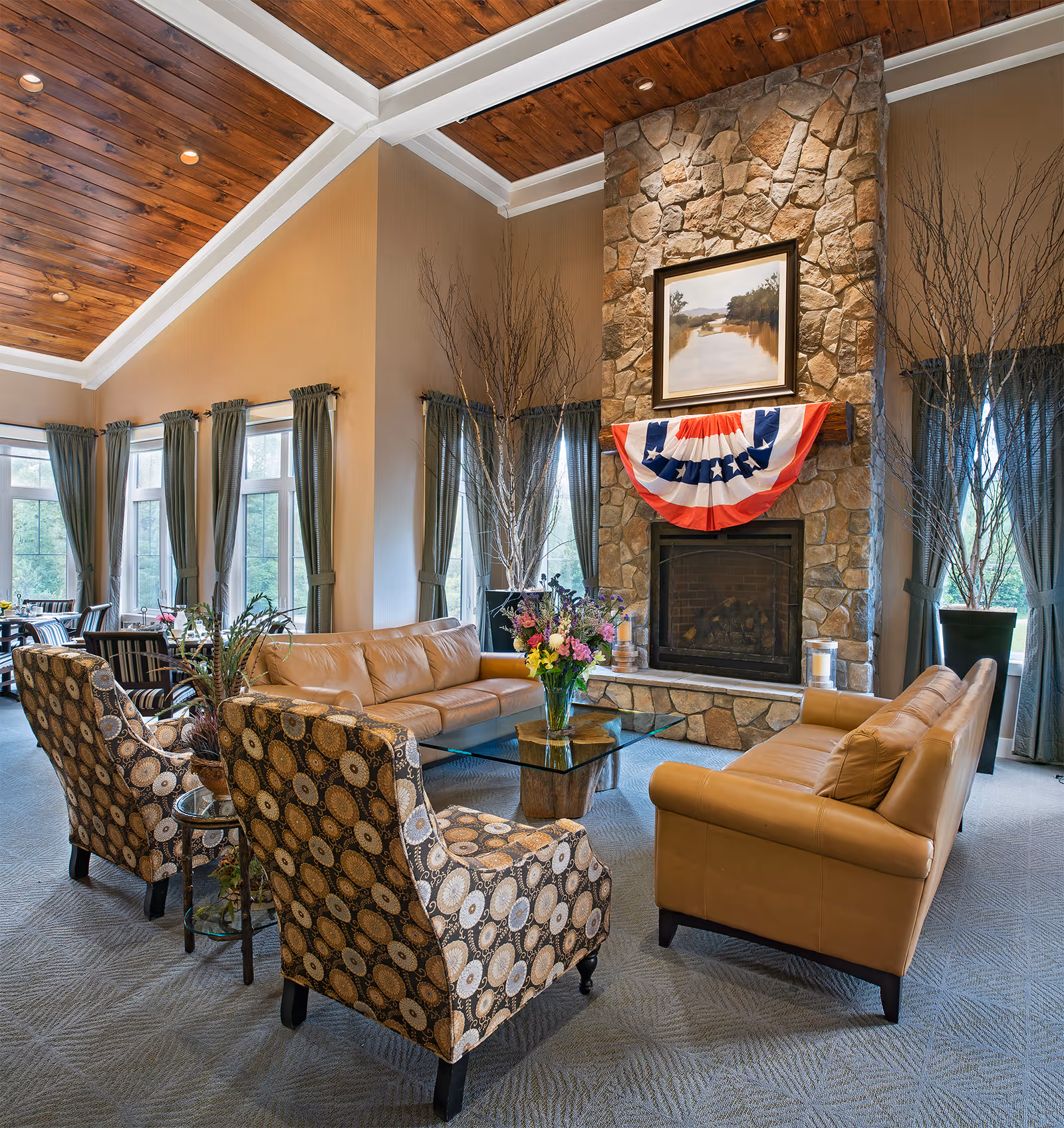 Bright communal living room with leather sofas and patterned armchairs arranged around a stone fireplace draped with a patriotic bunting.