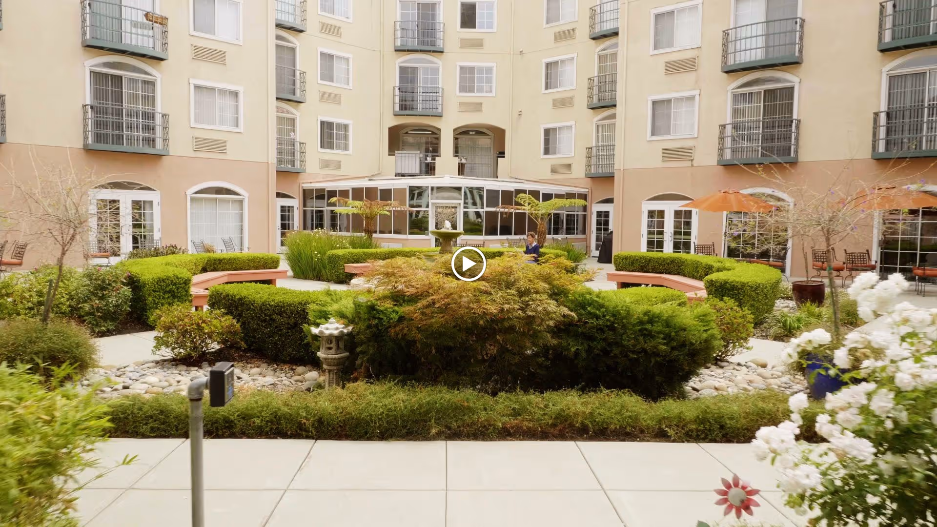 Outdoor courtyard area of a senior living facility with well-maintained bushes, trees, and flowers. There are curved benches around the garden and patio tables with umbrellas. The building surrounds the courtyard with multiple windows and balconies visible.