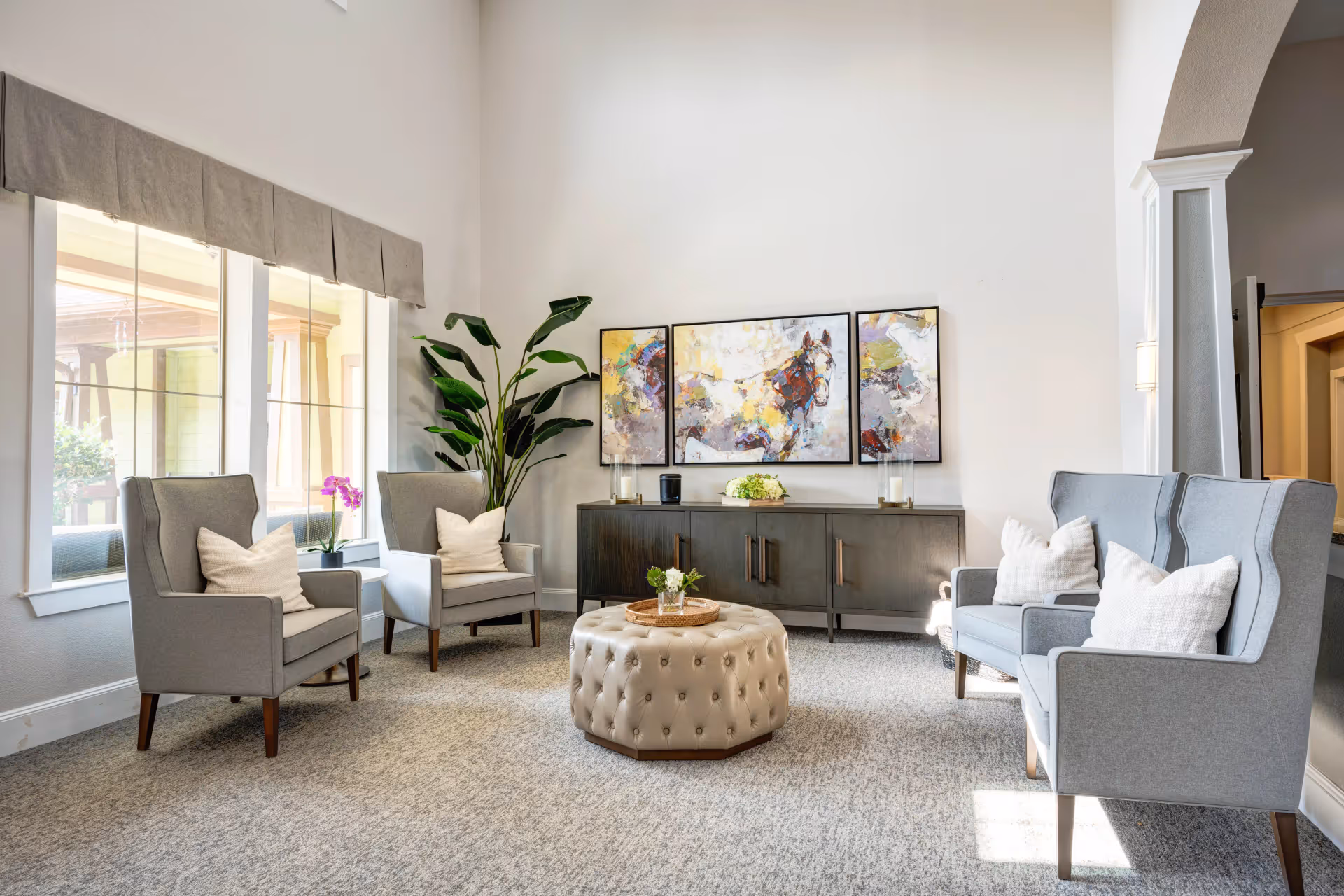 Bright assisted living lounge with gray armchairs arranged around a tufted ottoman, a sideboard with artwork and a large potted plant.