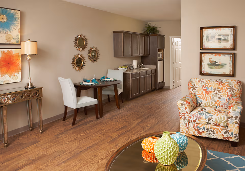 Bright furnished living room featuring a patterned armchair, a coffee table with colorful vases, a small dining table, and a kitchenette against the wall.