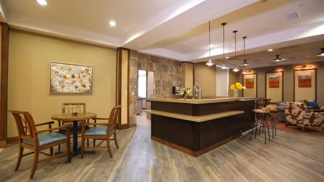Interior view of a common area in a senior living facility featuring a wooden round table with four chairs on the left, a large dark wood reception or bar counter in the center with pendant lights hanging above, and a seating area with patterned armchairs and framed artwork on the far right. The walls are painted beige and the floor has wood-like flooring.