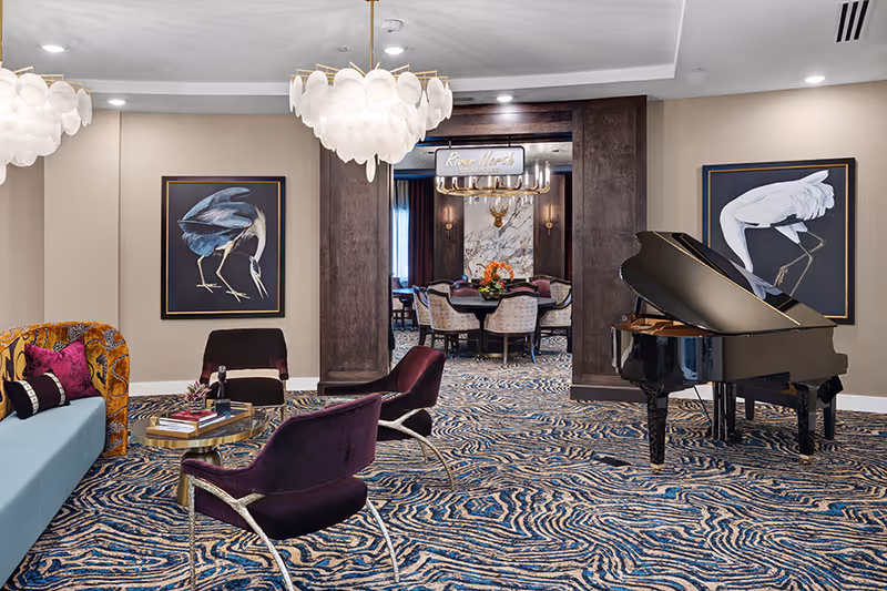 A stylish interior lounge area featuring a blue sofa with colorful pillows, two purple velvet chairs, a glass coffee table with books and decor, a black grand piano, and two framed bird paintings on the walls. In the background, a dining area with a round table and chairs is visible through a dark wood-framed doorway, with a sign above reading 'River North Steakhouse'. The room has patterned carpet and modern pendant lighting.