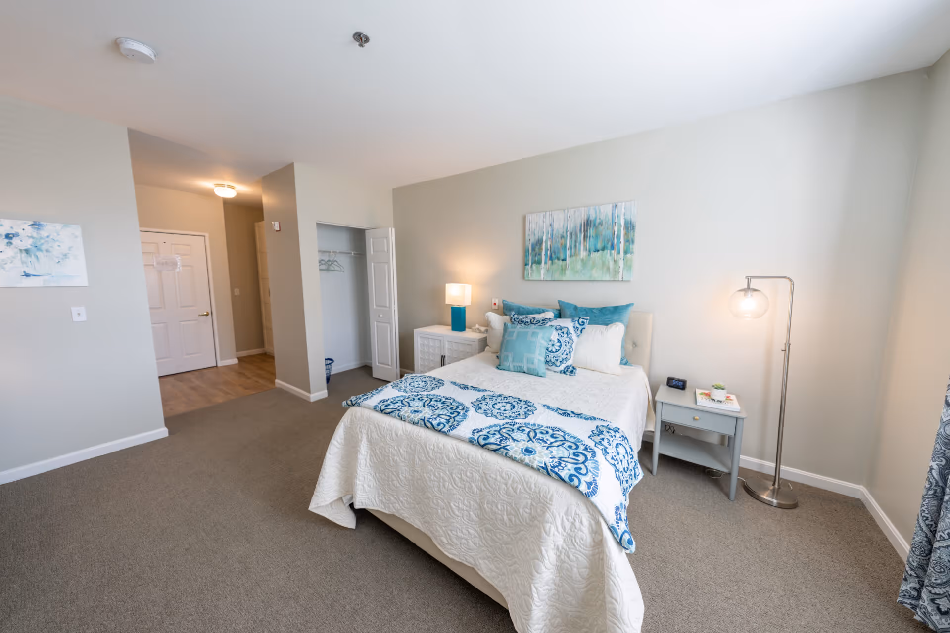 A neatly arranged bedroom with a bed covered in white and blue patterned bedding, two bedside tables with lamps, a small open closet with hangers, a painting above the bed, and a door leading to another room.