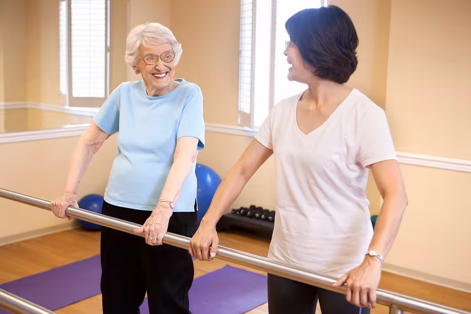 An elderly woman and a younger woman holding onto a ballet barre in a fitness or exercise room, smiling and looking at each other. The room has wooden floors, exercise mats, and exercise balls in the background.