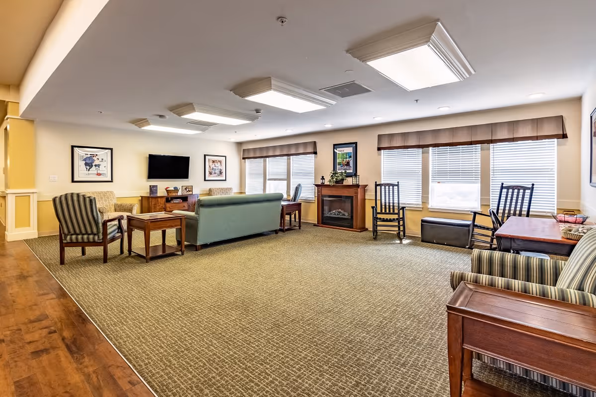 A spacious living room area in Willowbrook Place featuring a green sofa, multiple armchairs, a wooden coffee table, a flat-screen TV mounted on the wall, a fireplace with framed artwork above it, and large windows with blinds and valances allowing natural light to fill the room.
