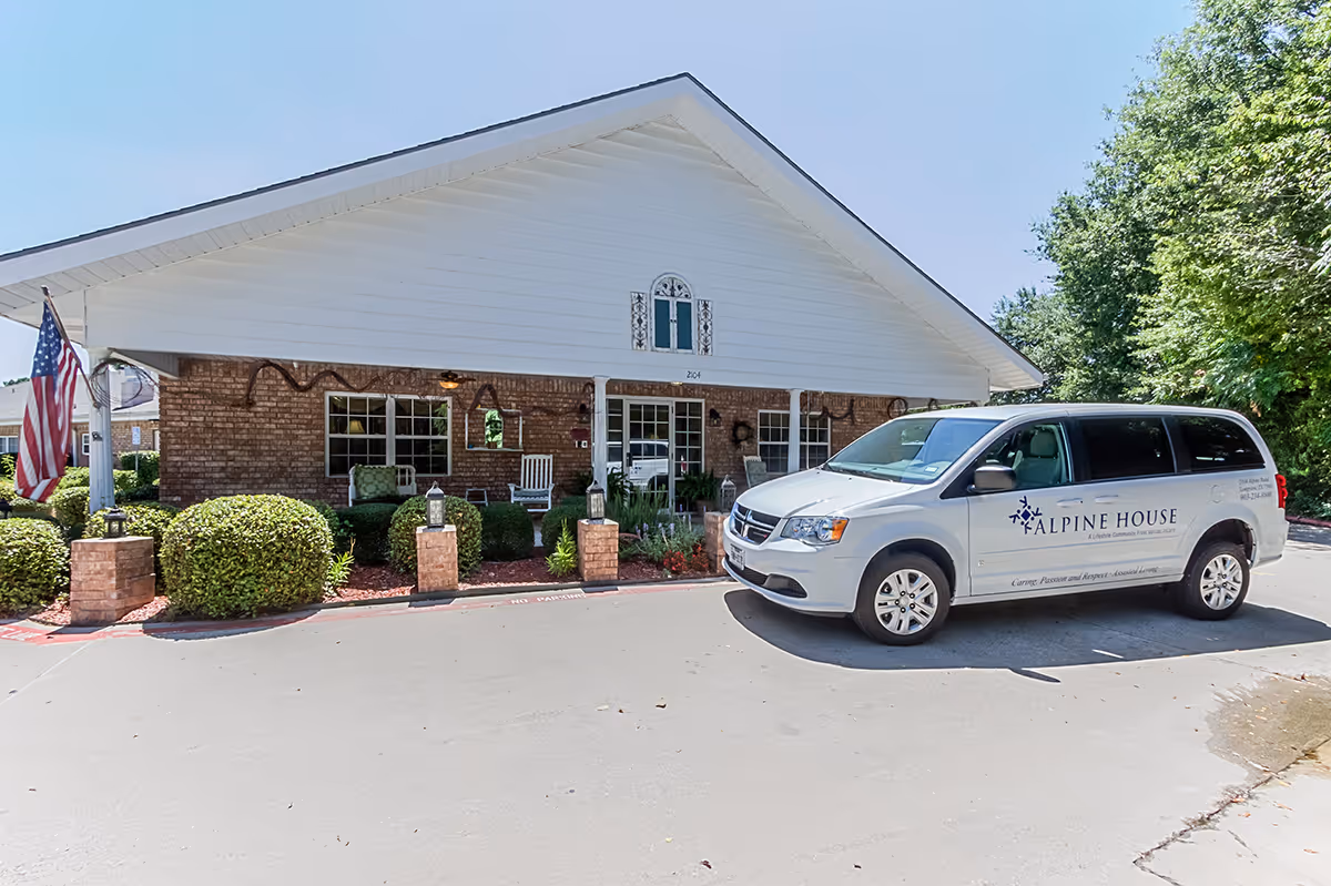 Exterior view of Alpine House facility showing a brick building with a white triangular roof. There is a white van parked in front with the Alpine House logo and text on its side. The area is landscaped with bushes and an American flag is displayed near the entrance.