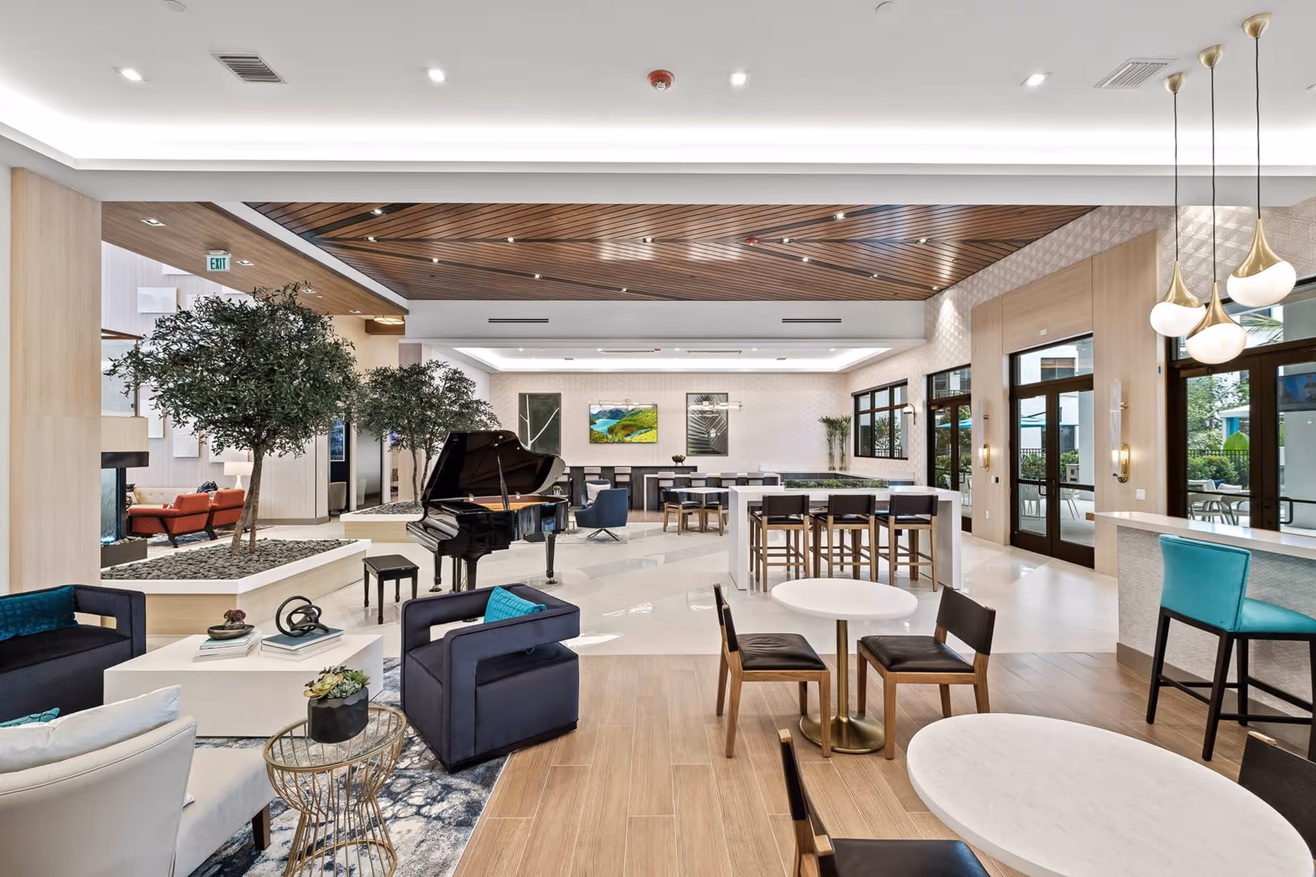 Spacious and modern common area in The Residences at Monterra Commons featuring a black grand piano, multiple seating arrangements with chairs and tables, indoor trees, large windows, and stylish pendant lighting.