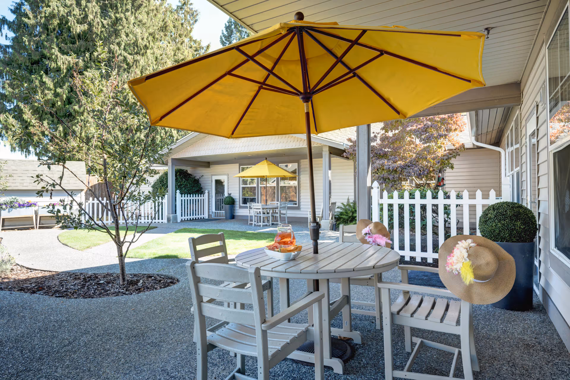 Outdoor patio area at a senior living facility with a round table and four chairs under a large yellow umbrella. Two of the chairs have straw hats decorated with flowers hanging on their backs. In the background, there is a white picket fence, a small tree, and another table with a yellow umbrella. The area is surrounded by greenery and buildings with beige siding.