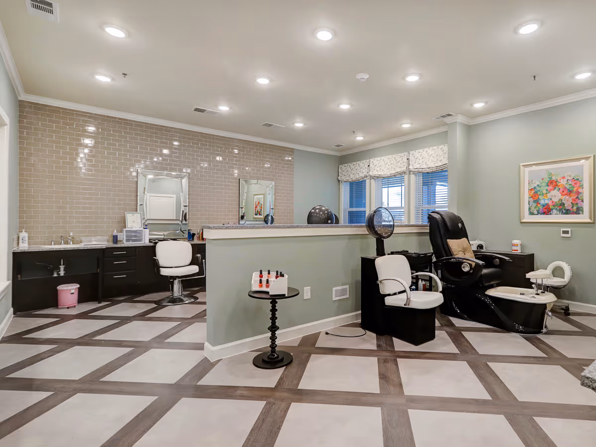 Interior view of a beauty salon area in a senior living facility with salon chairs, mirrors, a pedicure station, nail polish display, and a floral painting on the wall. The room has a tiled floor with a crisscross pattern and recessed ceiling lights.