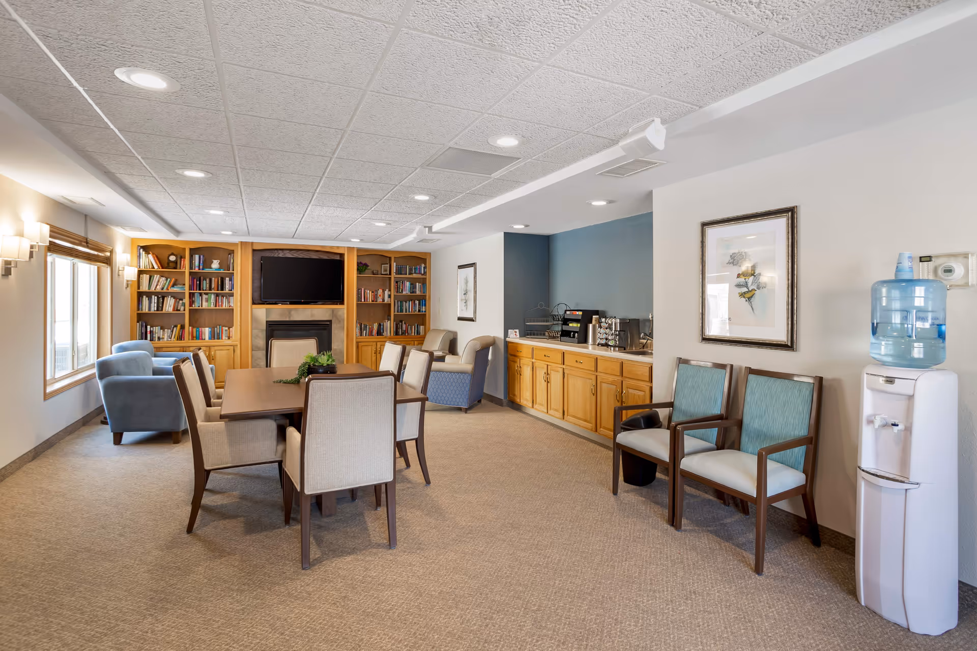 A senior living facility common area with a rectangular wooden table surrounded by beige upholstered chairs in the center. To the left, there are blue armchairs near a window with wooden blinds. The back wall features built-in wooden bookshelves filled with books and a flat-screen TV mounted above a fireplace. On the right side, there is a wooden cabinet with a coffee station and a water cooler next to two wooden chairs with teal cushions. The room has beige carpet and a white textured ceiling with recessed lighting.