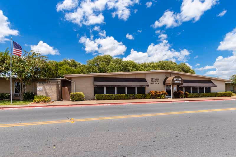 Exterior view of a single-story building with beige walls and black awnings over the windows. The building has a sign that reads 'Ketama Manor' above the entrance. There is an American flag on a flagpole to the left, some bushes and trees around the building, and a clear blue sky with scattered clouds.