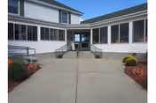 Entrance to a nursing home building with a concrete walkway leading to double glass doors. The building is white with large windows on either side of the entrance. There are Halloween decorations including carved pumpkins and potted flowers on both sides of the walkway.