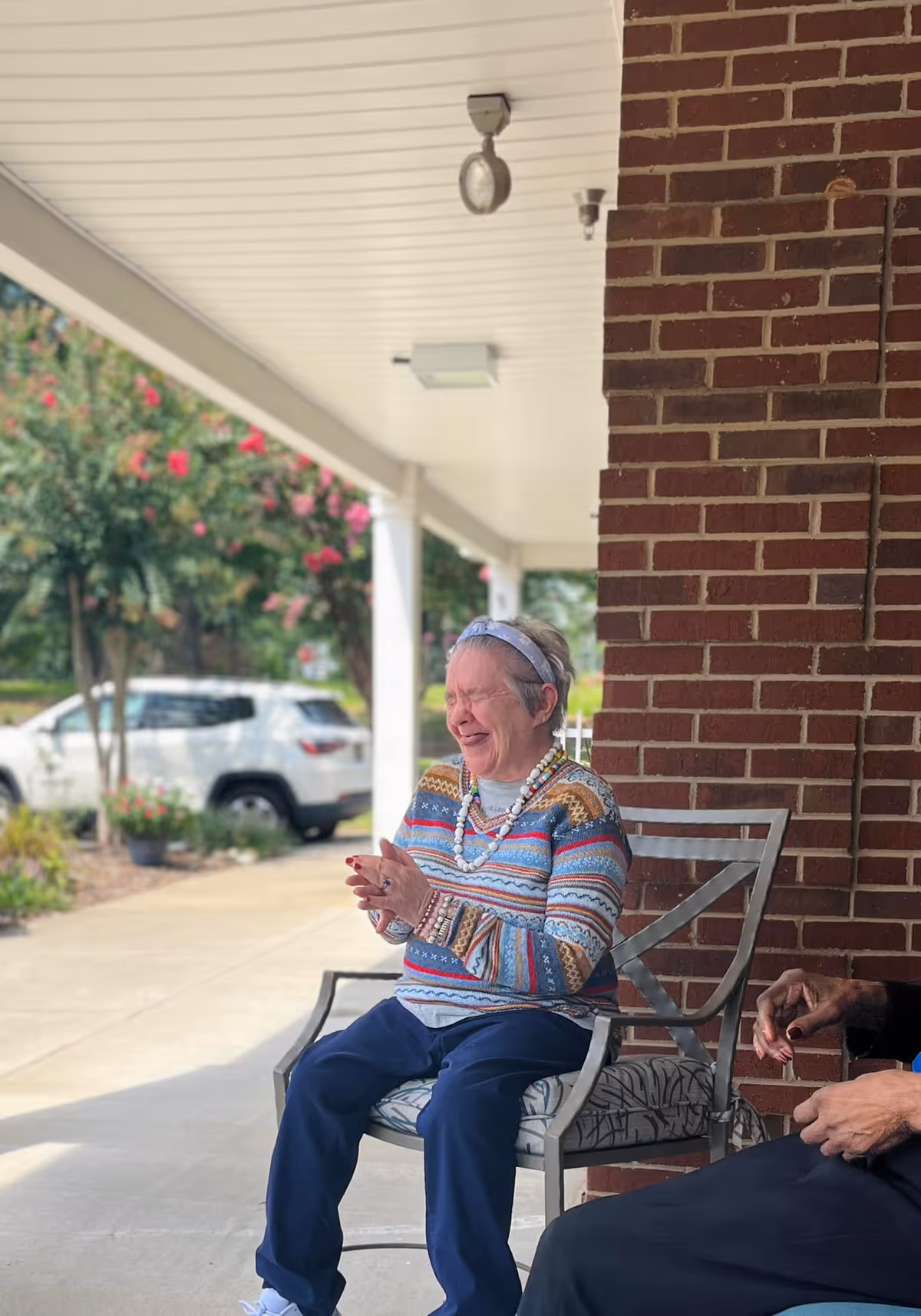 An elderly woman wearing a colorful striped sweater and a headband is sitting on a cushioned metal chair on a covered porch outside a brick building. She is smiling and clapping her hands. Another person is partially visible sitting next to her. In the background, there are trees, flowers, and a white SUV parked on a driveway.