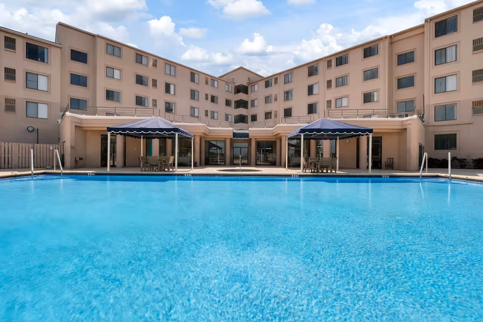 Outdoor swimming pool in front of a multi-story senior living facility building with beige exterior walls. Two shaded seating areas with tables and chairs are visible near the poolside. The sky is partly cloudy.