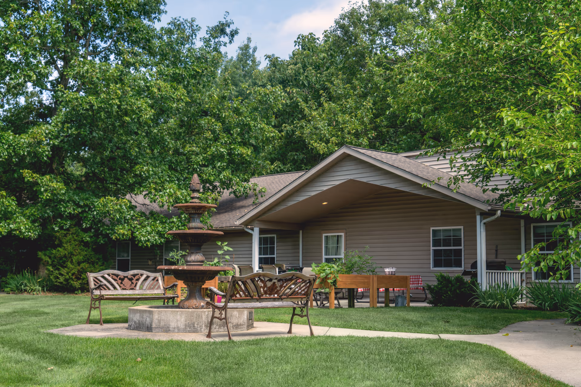 Outdoor area of a senior living facility with a three-tiered fountain surrounded by metal benches on a concrete platform, green grass, trees, and a single-story building with a covered porch in the background.