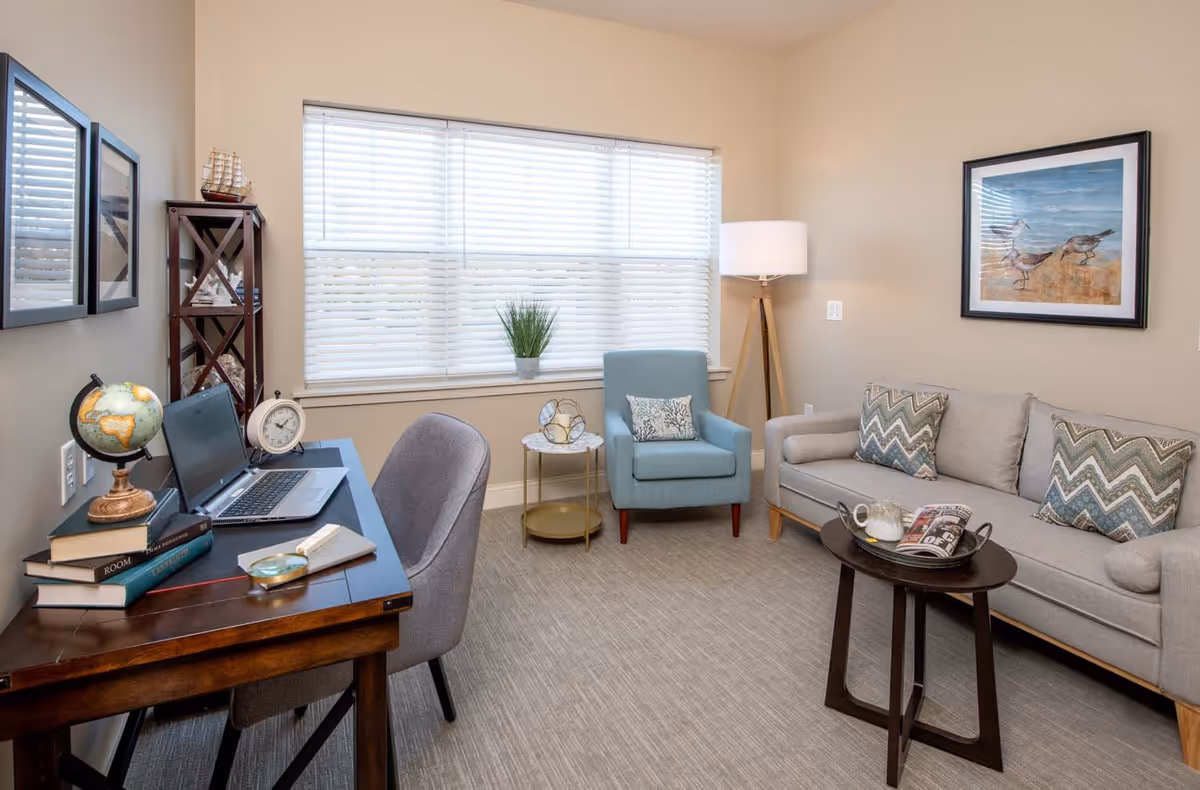 A cozy living room with a beige sofa adorned with patterned pillows, a light blue armchair with a decorative pillow, a round wooden coffee table holding a teapot and a magazine, a wooden desk with a laptop, books, a globe, and a clock, a gray upholstered chair, a tall floor lamp, and a window with white blinds letting in natural light. The walls are decorated with framed artwork and the carpet is light gray.