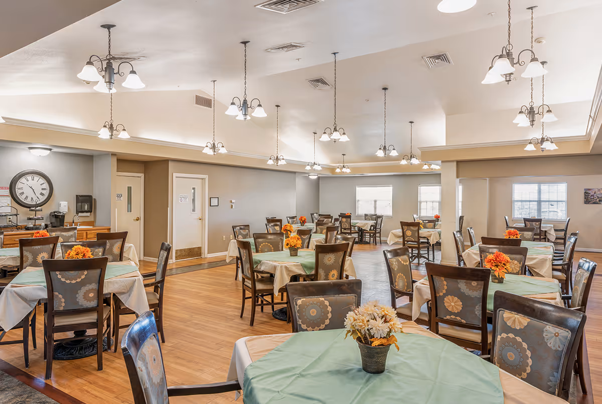 A spacious dining room in a senior living facility with multiple round tables covered with light green and beige tablecloths. Each table has a floral centerpiece with orange and white flowers. The room has wooden flooring, beige walls, multiple windows allowing natural light, and several hanging light fixtures on the ceiling. Chairs with patterned upholstery surround the tables.