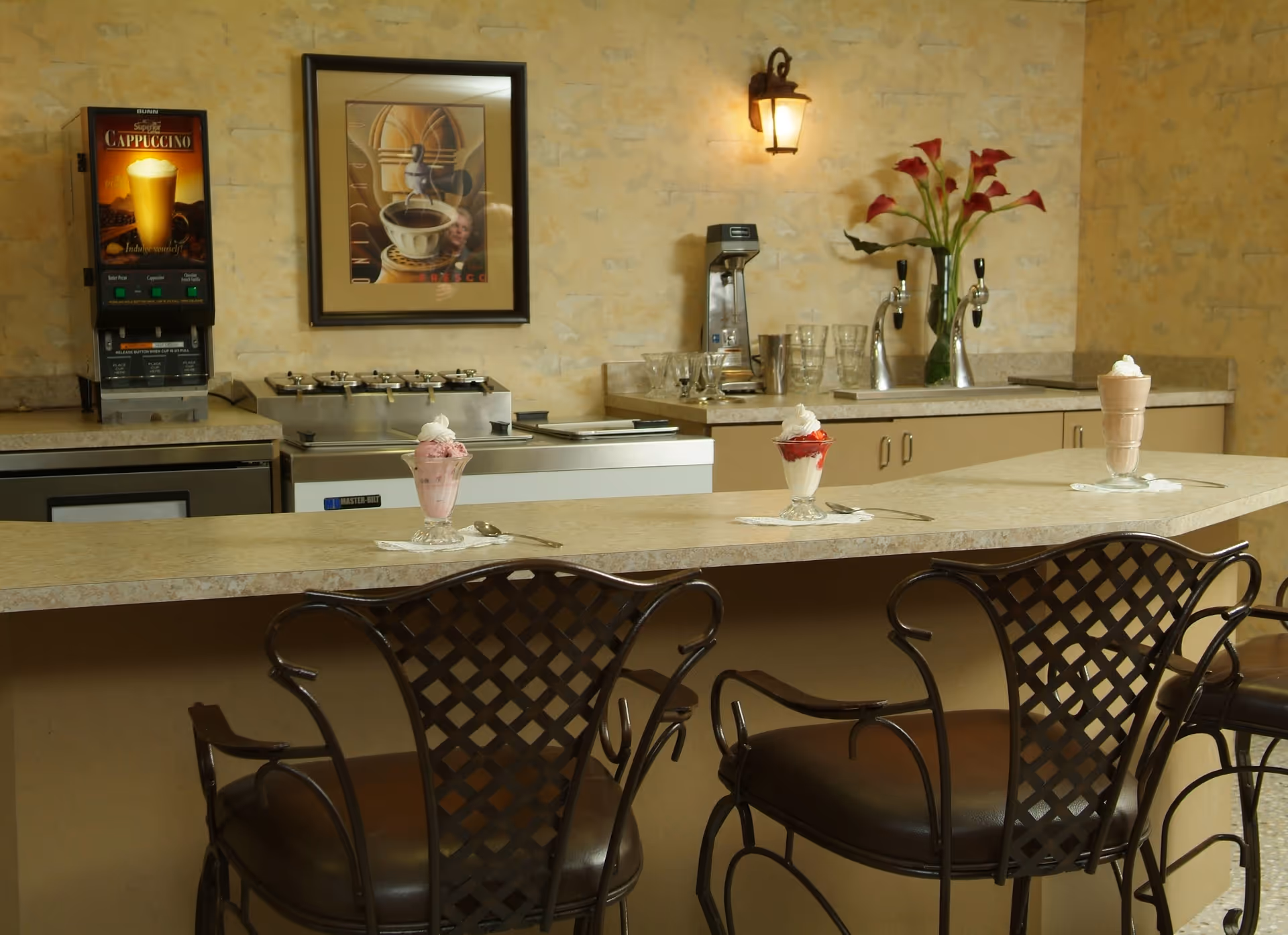 Service counter with three stools and three ice cream sundaes, coffee machines and framed art on the wall in a dining/kitchen area.