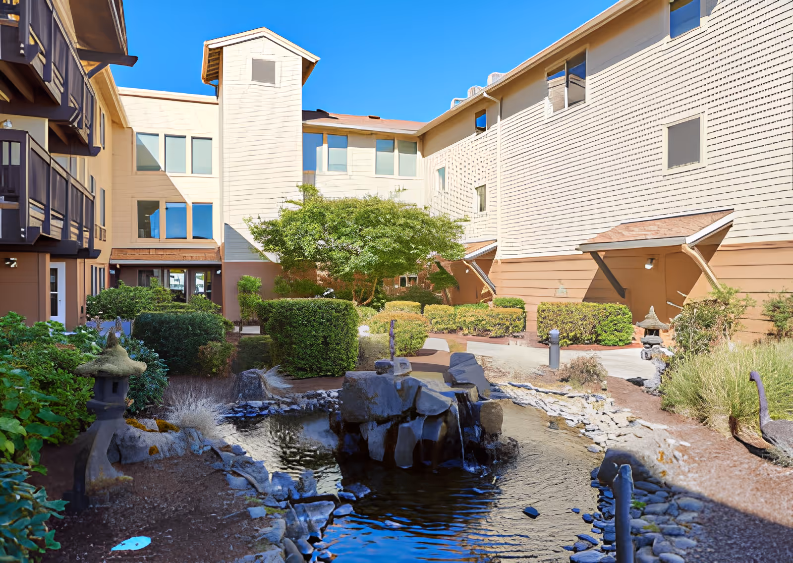 Sunlit courtyard with a rock-lined pond and fountain surrounded by shrubs and a multi-story residence building.