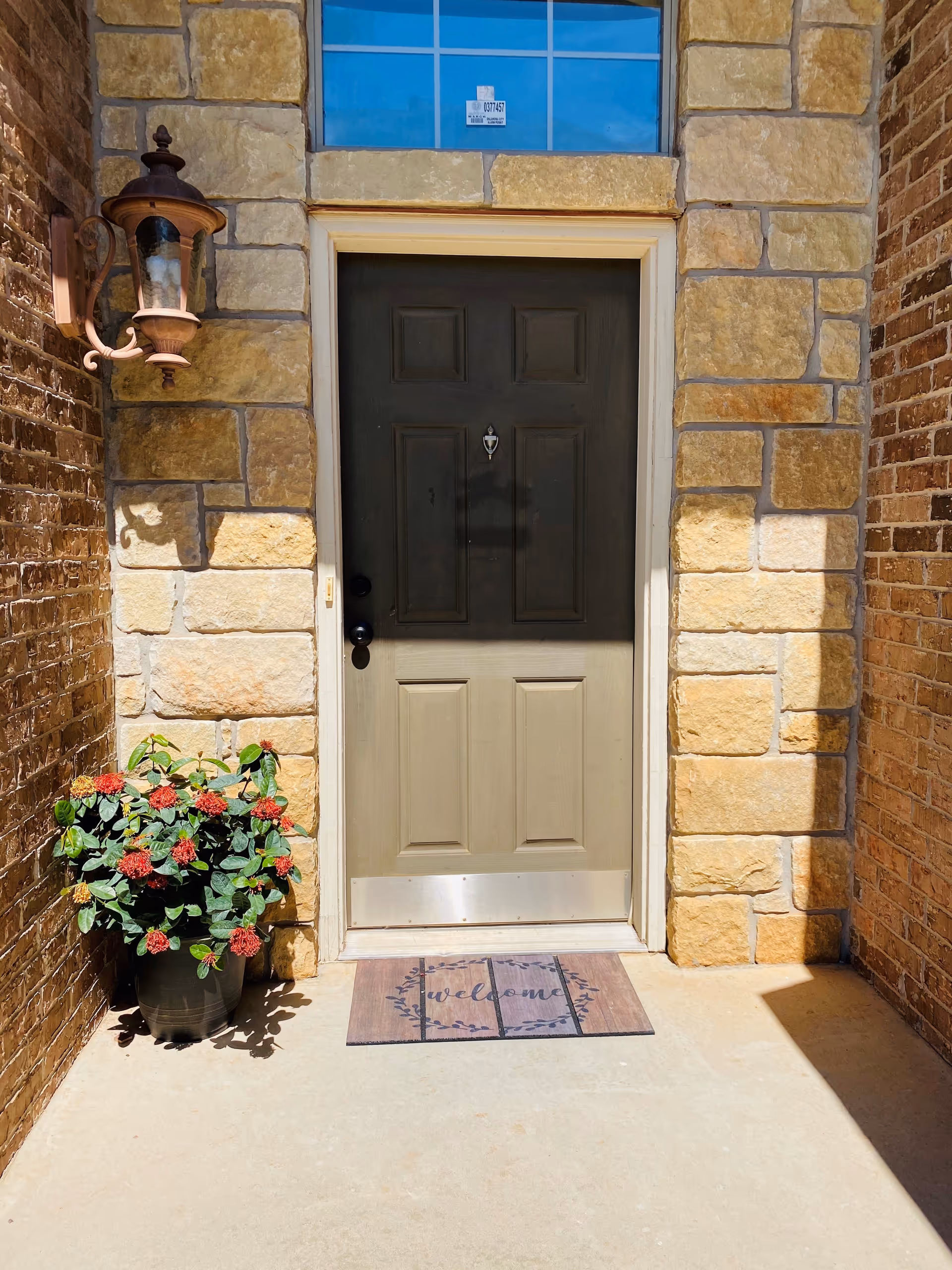 Front entrance door of a building with a stone and brick exterior. There is a black door with a silver kick plate and a small window above it. A wall-mounted lantern light fixture is on the left side, and a potted plant with red flowers is placed on the ground next to the door. A welcome mat is laid out in front of the door.