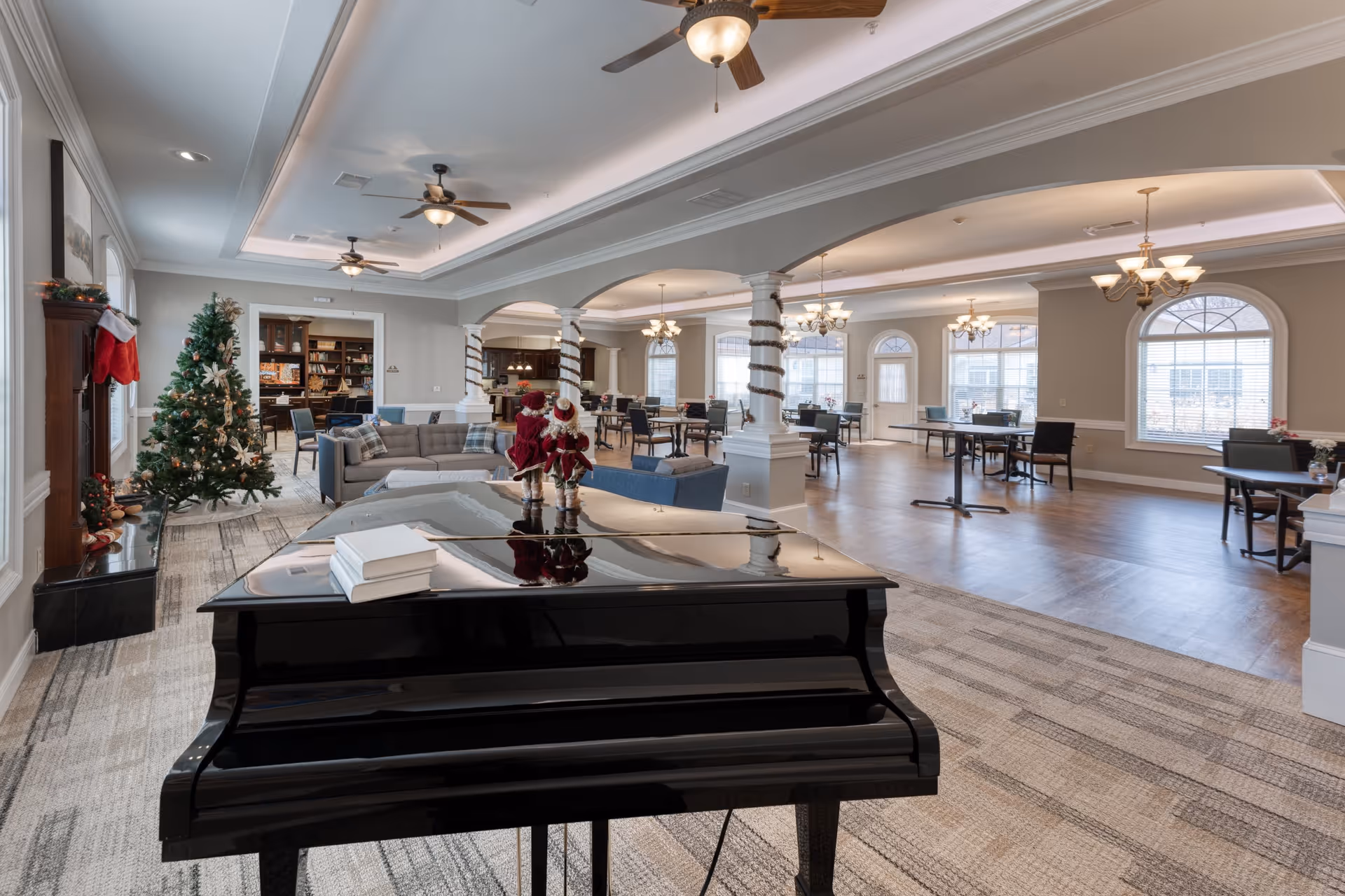 Spacious and well-lit senior living common area decorated for Christmas with a grand piano in the foreground, a Christmas tree and stockings on the left, comfortable seating areas, and multiple dining tables with chairs under ceiling fans and chandeliers.