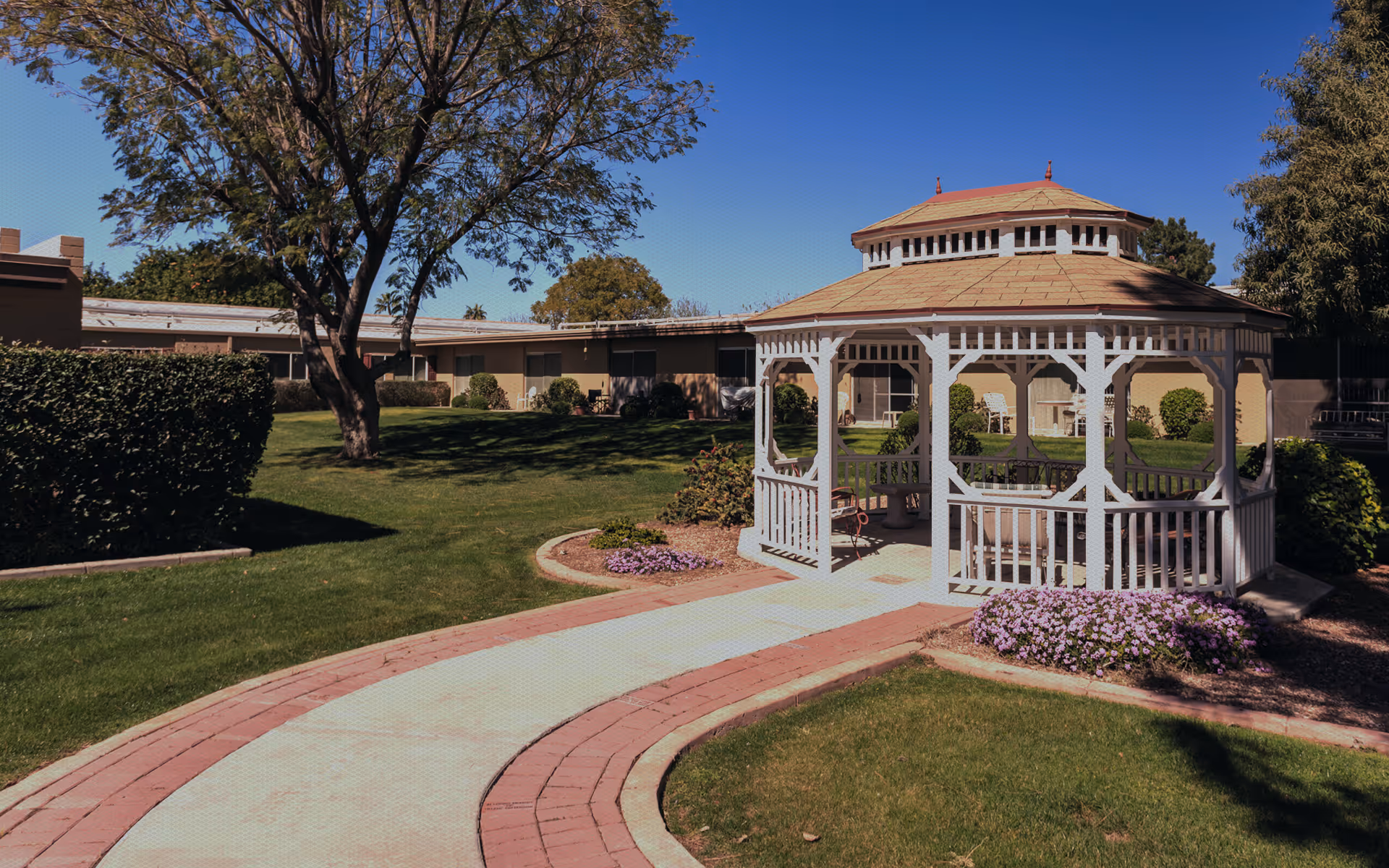 A sunny outdoor garden area at a senior living facility featuring a white wooden gazebo with seating inside, surrounded by green grass, bushes, and purple flowers. A paved walkway curves towards the gazebo, and single-story buildings are visible in the background under a clear blue sky.