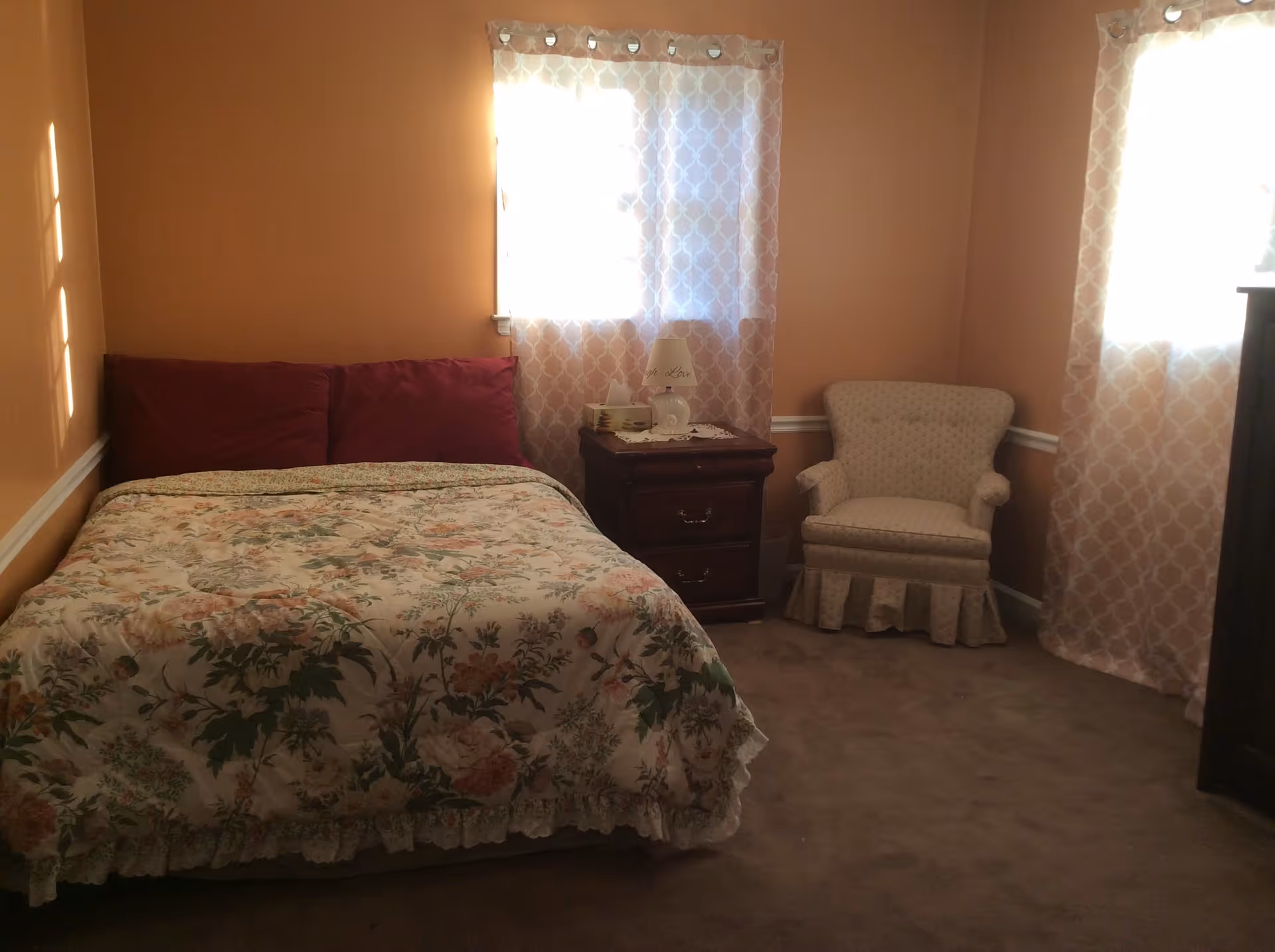 A cozy bedroom with peach-colored walls, a bed with floral bedding and red pillows, a wooden nightstand with a lamp and some items on top, a cushioned armchair with a skirted bottom, and two windows with patterned curtains letting in natural light.