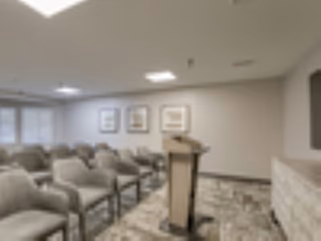 A meeting or presentation room with rows of beige upholstered chairs arranged facing a wooden podium. The room has neutral-colored walls with three framed pictures hanging on one wall, and a carpeted floor with a patterned design. The lighting is provided by ceiling fixtures.