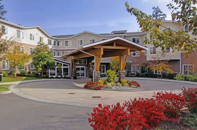 Exterior view of Radcliffe Place senior living facility showing a multi-story building with beige and brown siding, a covered entrance with wooden beams and stone pillars, surrounded by landscaped greenery and red bushes under a clear blue sky.