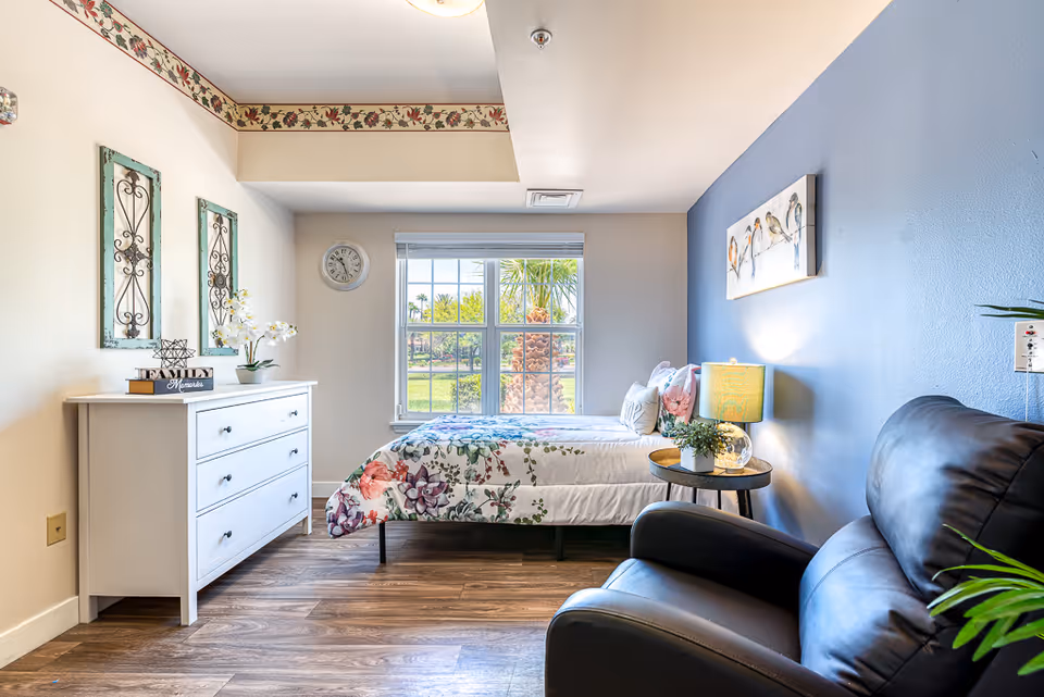 Bright, neatly decorated bedroom with a single bed by a window, white dresser, side table with a lamp, and a leather armchair.