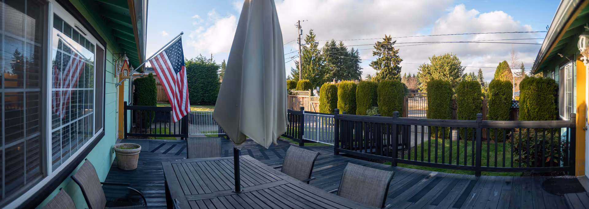 Outdoor patio area with a wooden table and chairs, a closed umbrella in the center, an American flag mounted on the green exterior wall of the building, and neatly trimmed bushes along the fence under a partly cloudy sky.