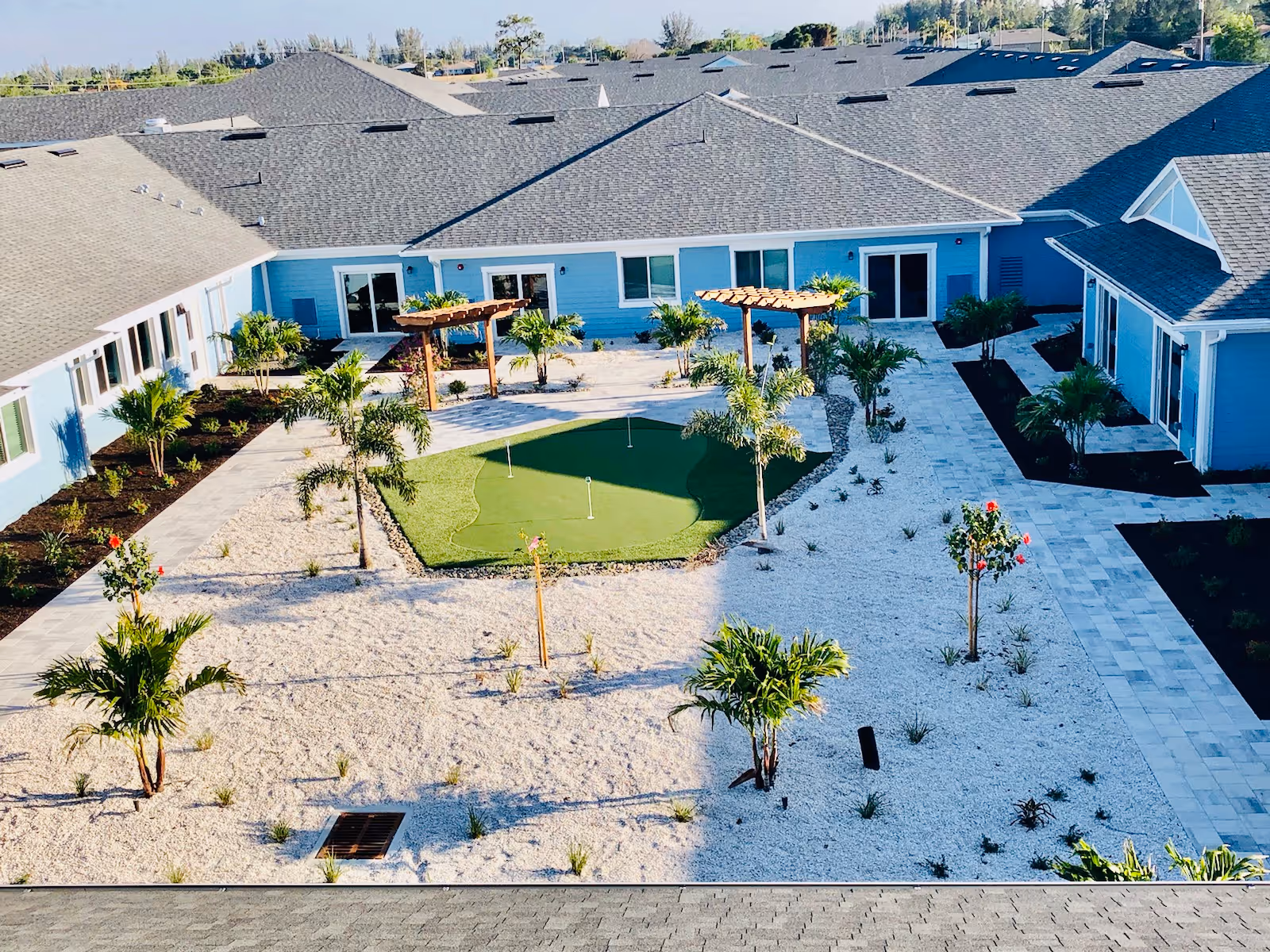 Aerial view of a sunny courtyard with a small putting green, pergolas, palm trees and walkways surrounded by a light-blue senior living building.